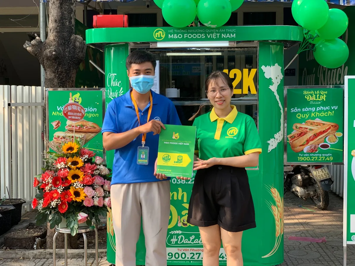 Hai người đứng trước quầy Dalabami và cầm tài liệu tư vấn nhượng quyền.