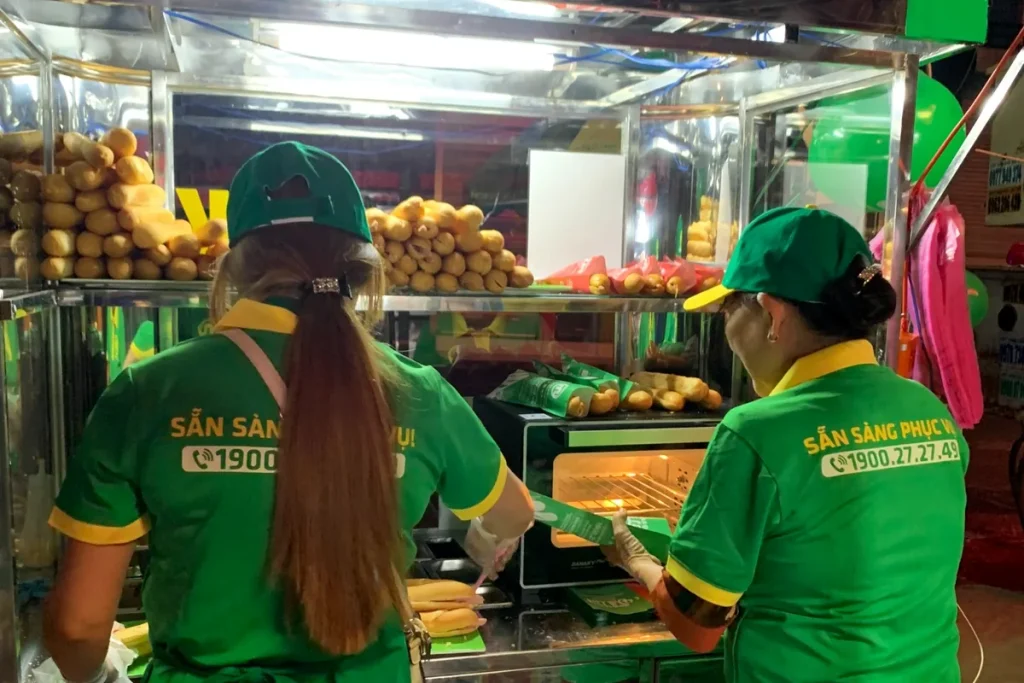 Hai nhân viên chuẩn bị bánh mì tại quầy kính có trưng bày bánh